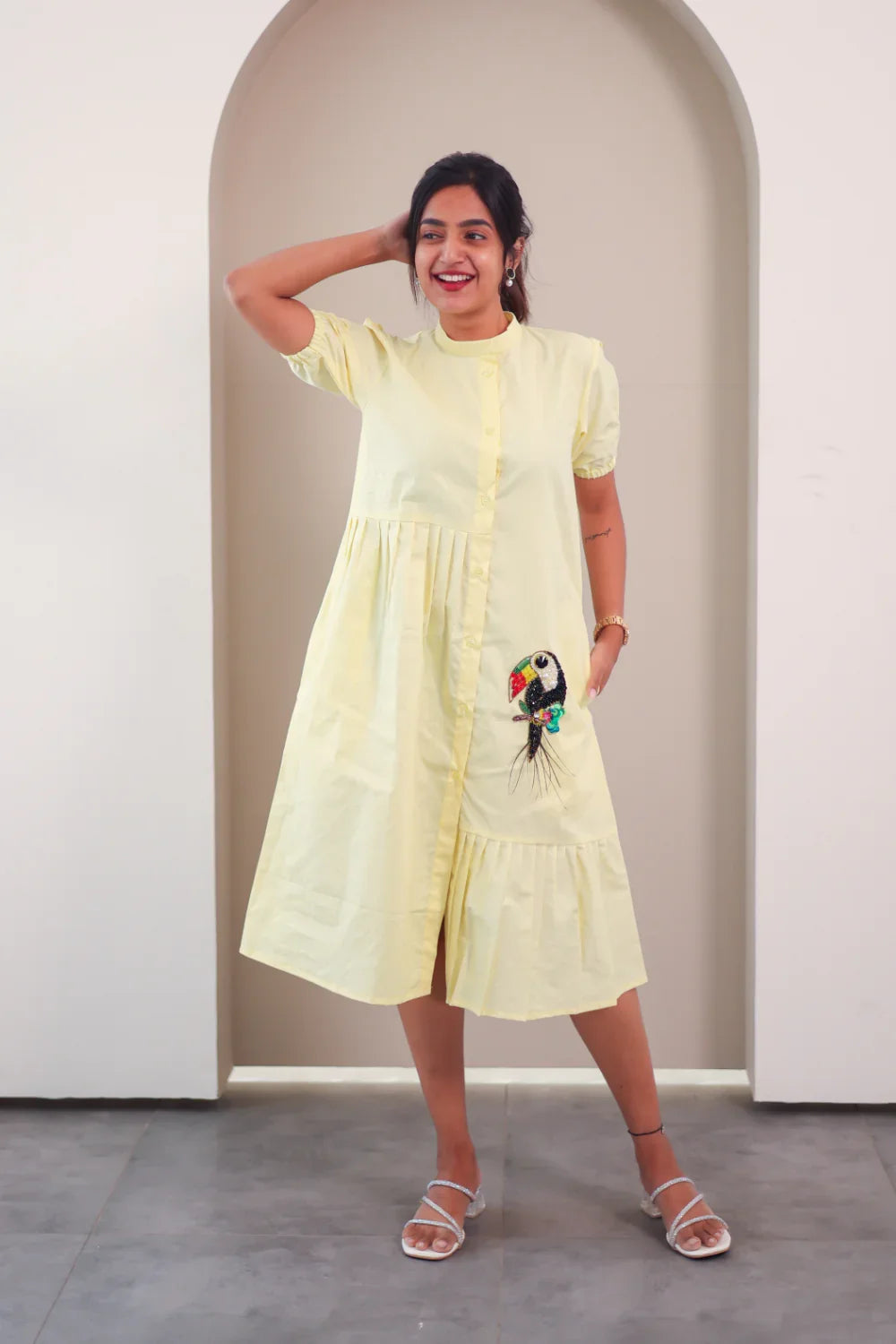Woman in yellow midi dress with parrot embroidery and short sleeves, standing indoors