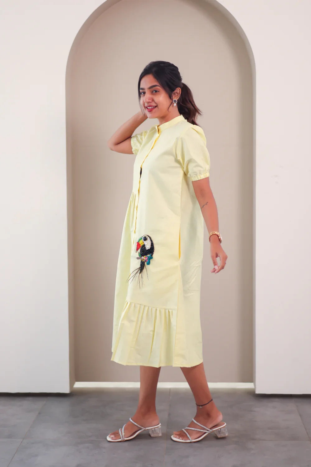 Woman in a light yellow midi dress with puff sleeves and toucan embroidery, posing indoors