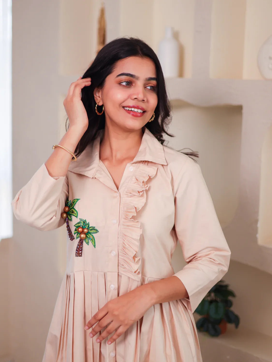 Woman wearing a beige dress with palm tree embroidery and ruffle detail, posing indoors