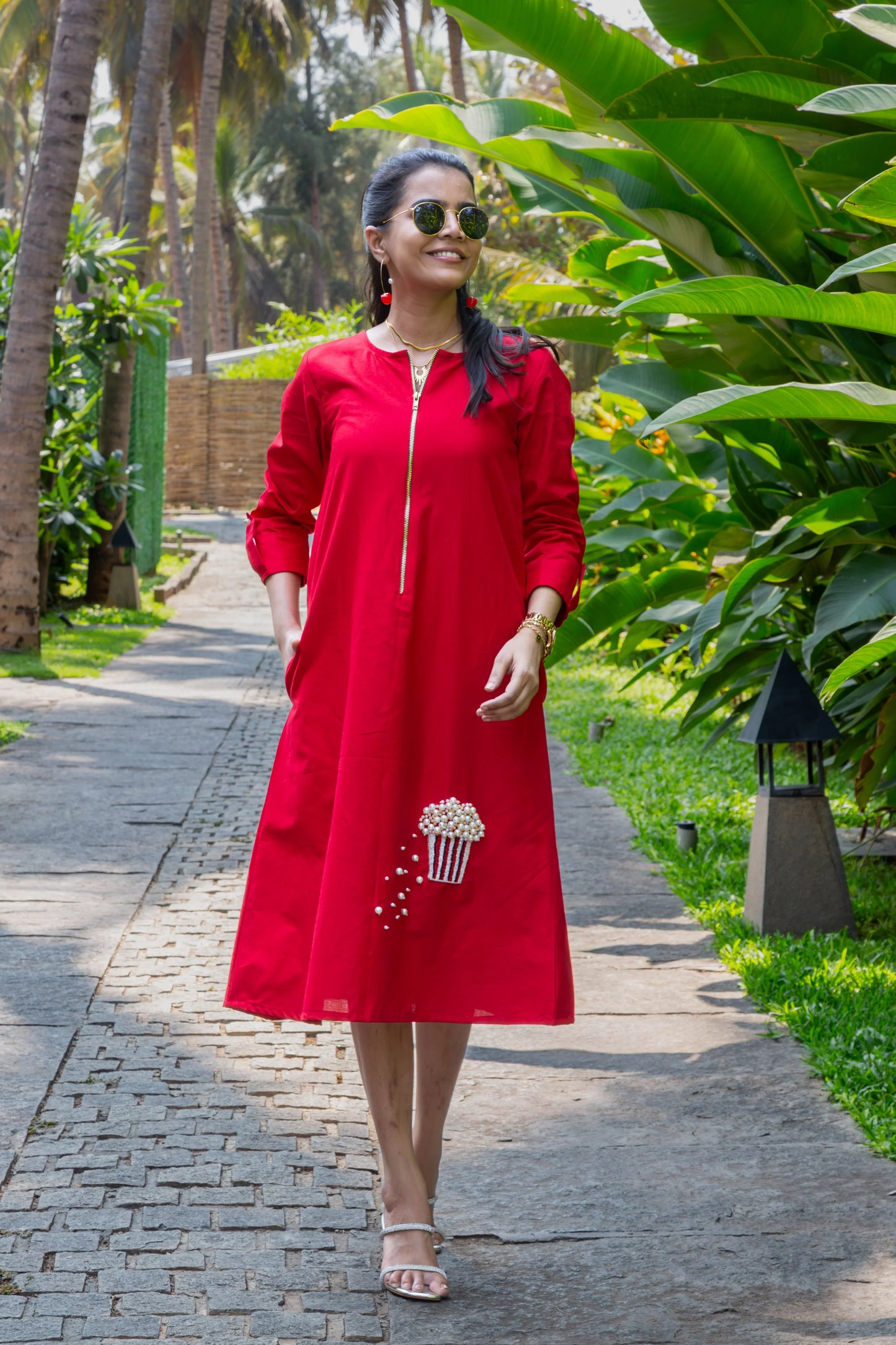 Woman in a red knee-length dress with popcorn embroidery, posing outdoors by green plants.