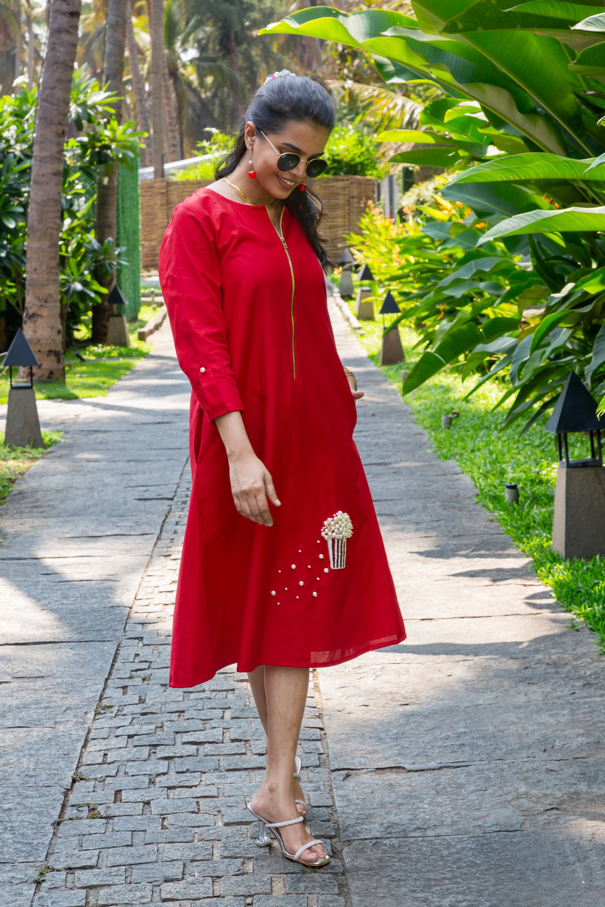 Woman in a red midi dress with pearl embellishments, walking outdoors on a sunny path
