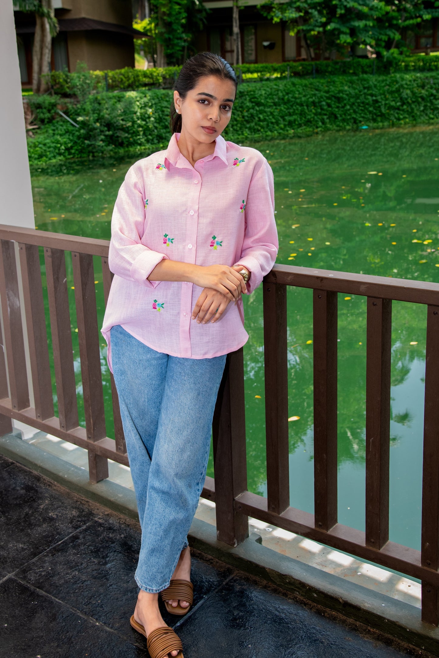 Woman in pink floral embroidered shirt with blue jeans, standing by garden pond, casual everyday style