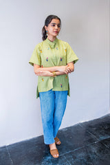 Woman wearing light green embroidered shirt, blue jeans, and sandals against plain wall