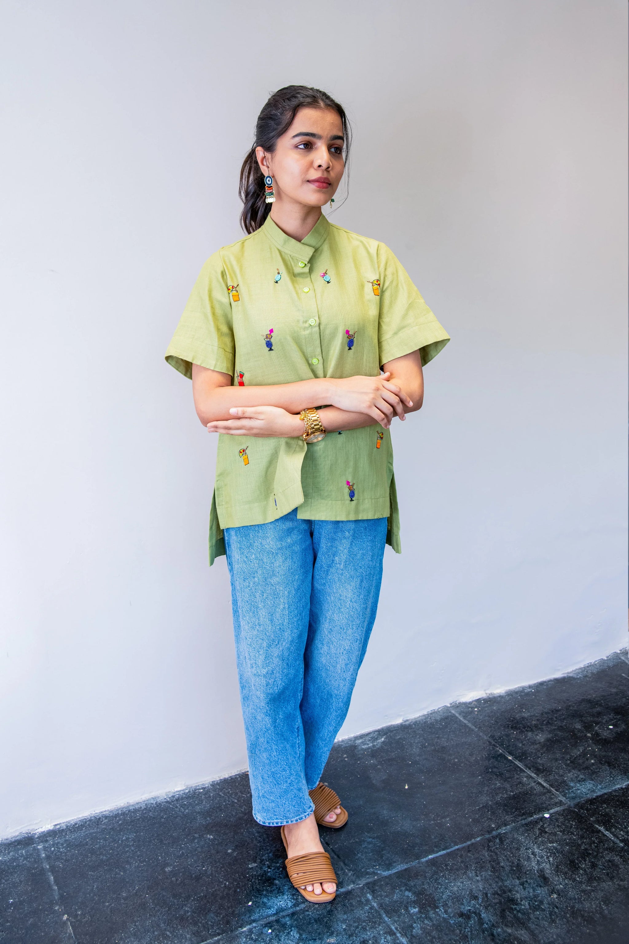 Woman wearing light green embroidered shirt, blue jeans, and sandals against plain wall