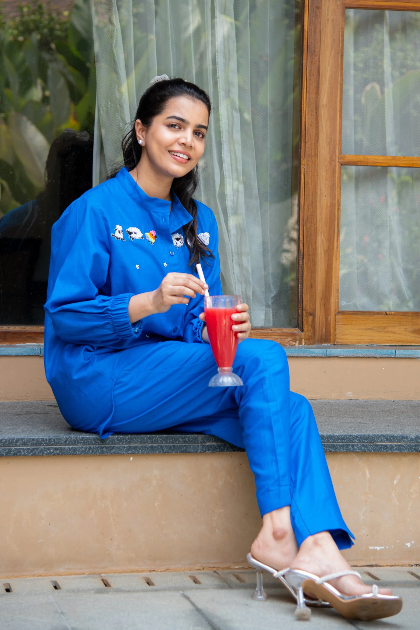 Woman in a blue co-ord set sitting outdoors, holding a glass of red juice, smiling