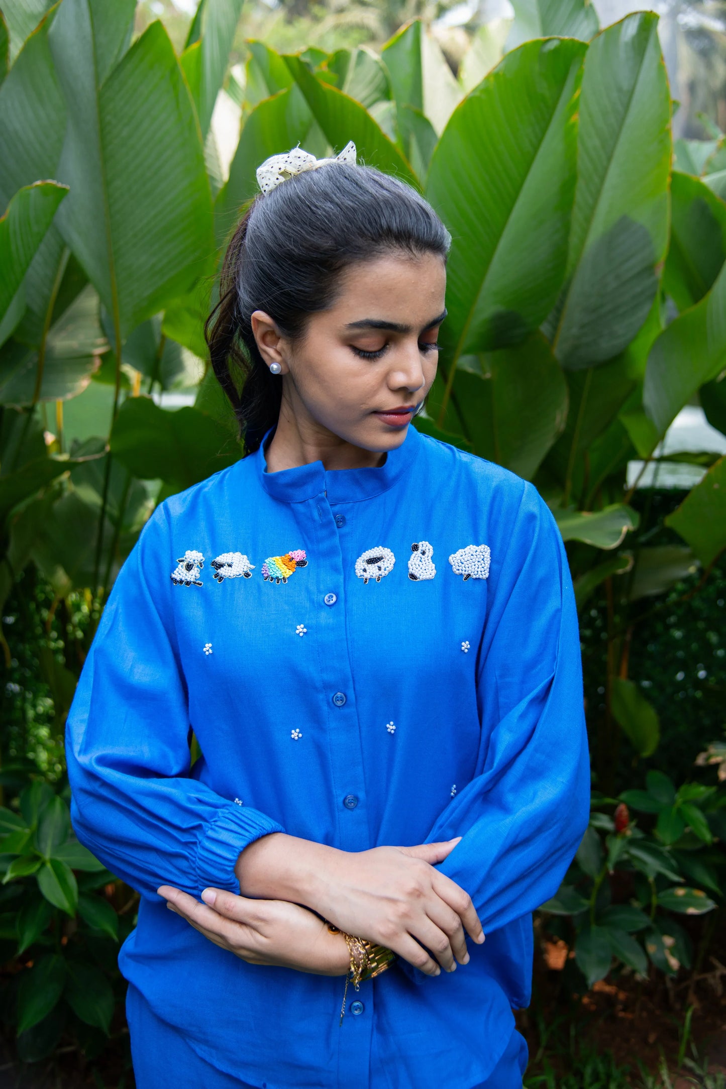 Woman in a blue shirt with sheep embroidery standing outdoors with green leaves