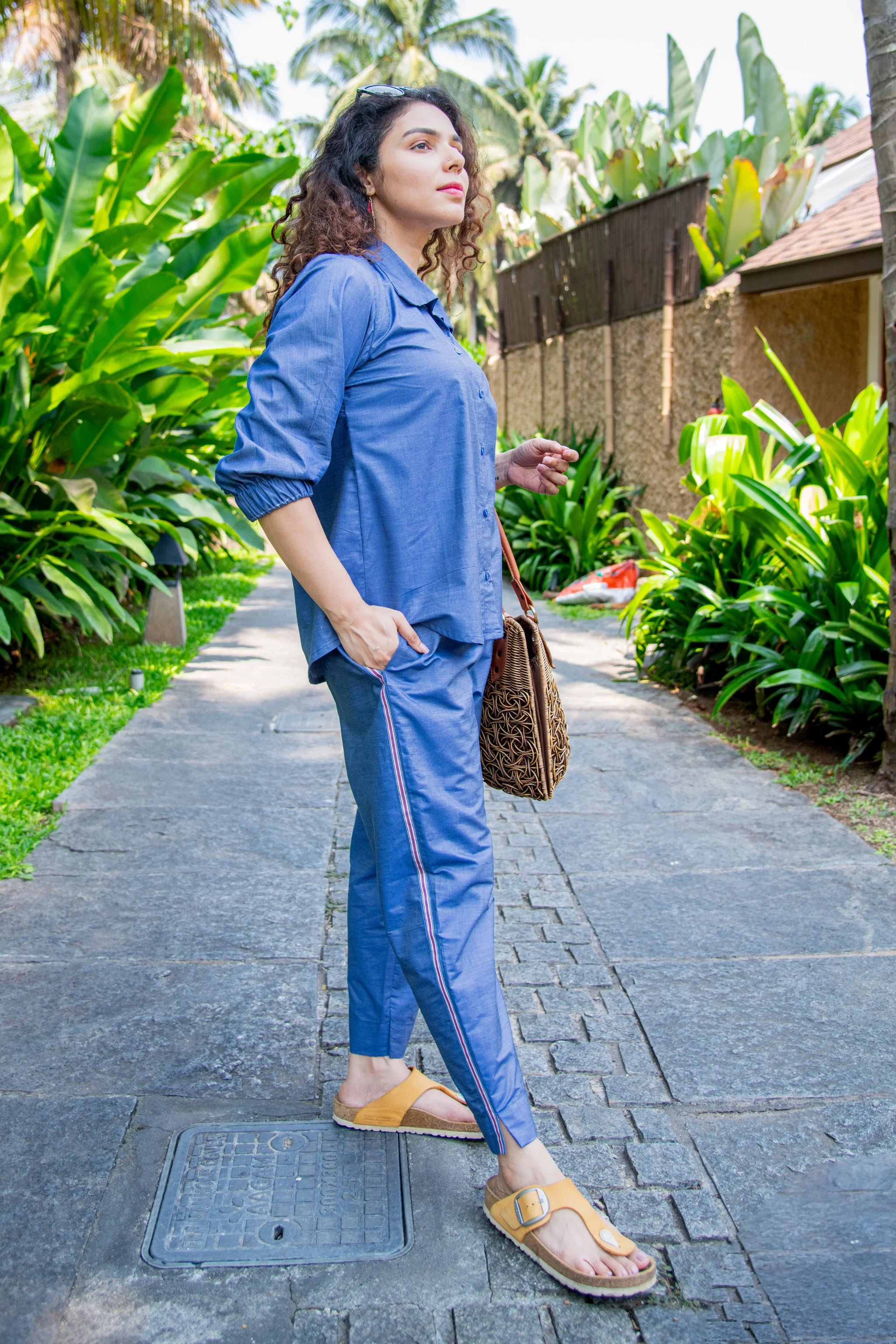 Woman in blue co-ord set with wicker bag and sandals, standing on a garden pathway