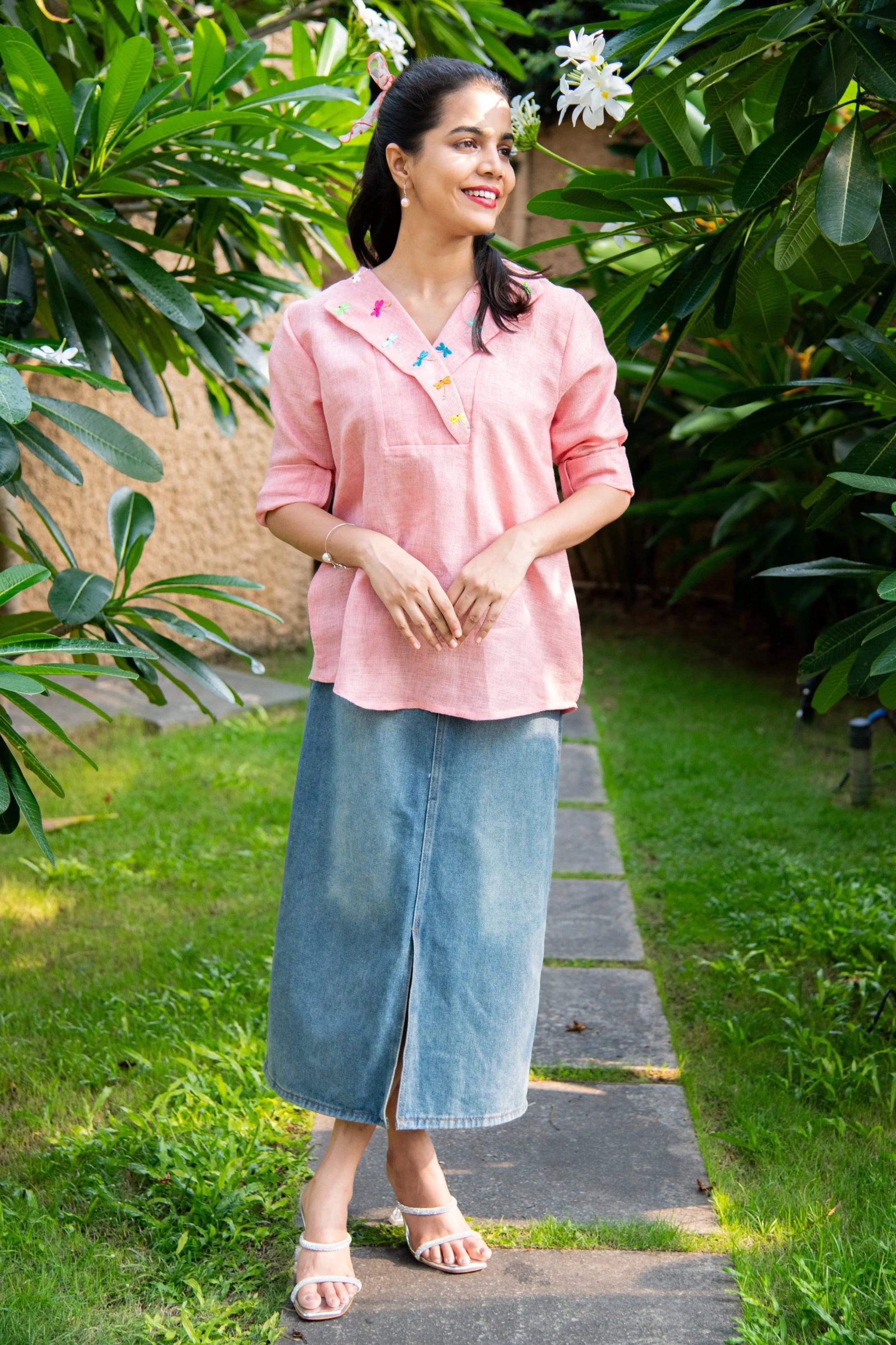 Woman in a pink embroidered top and denim skirt standing outdoors on a garden path