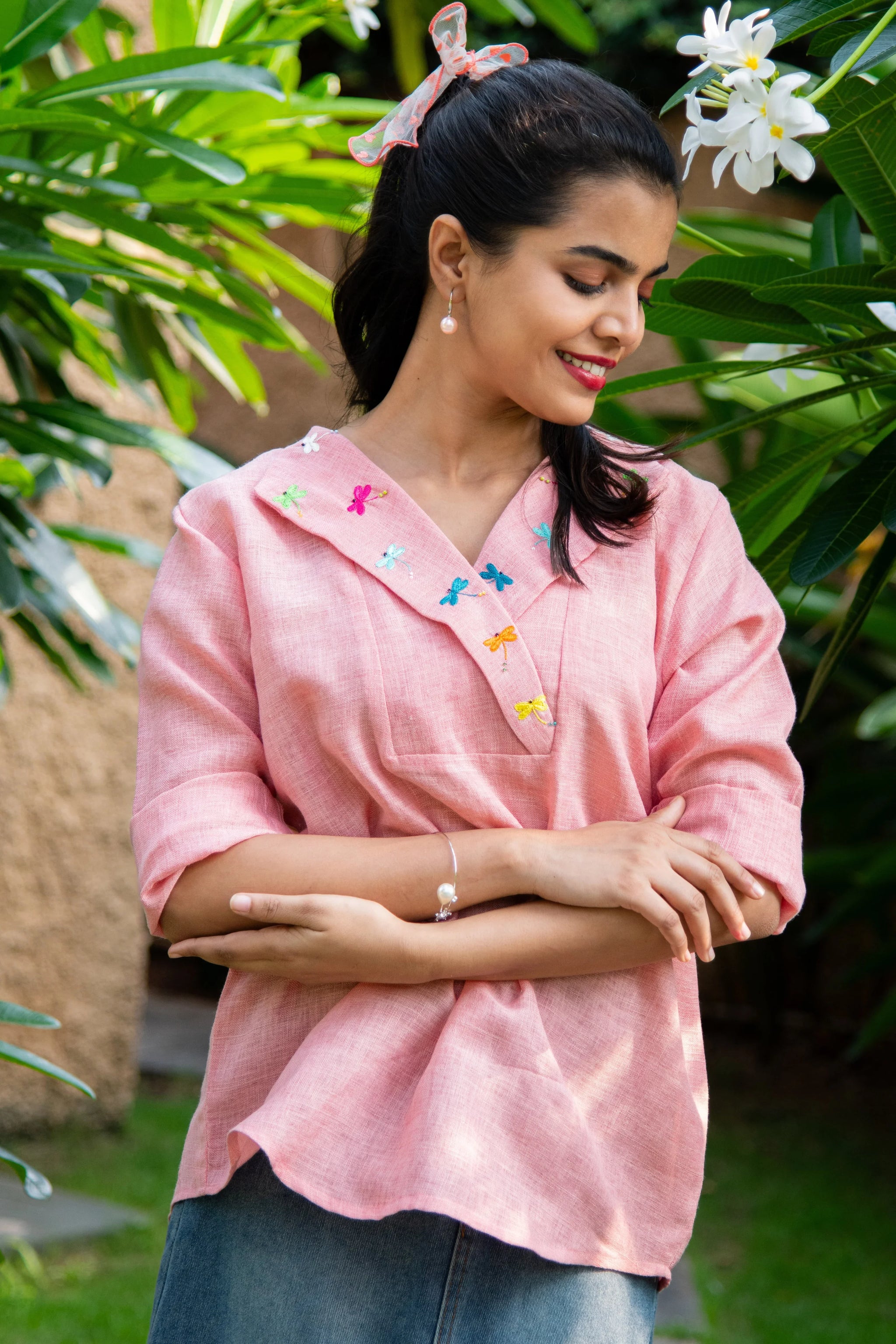 Woman in pink embroidered top with butterfly details, standing outdoors among green plants