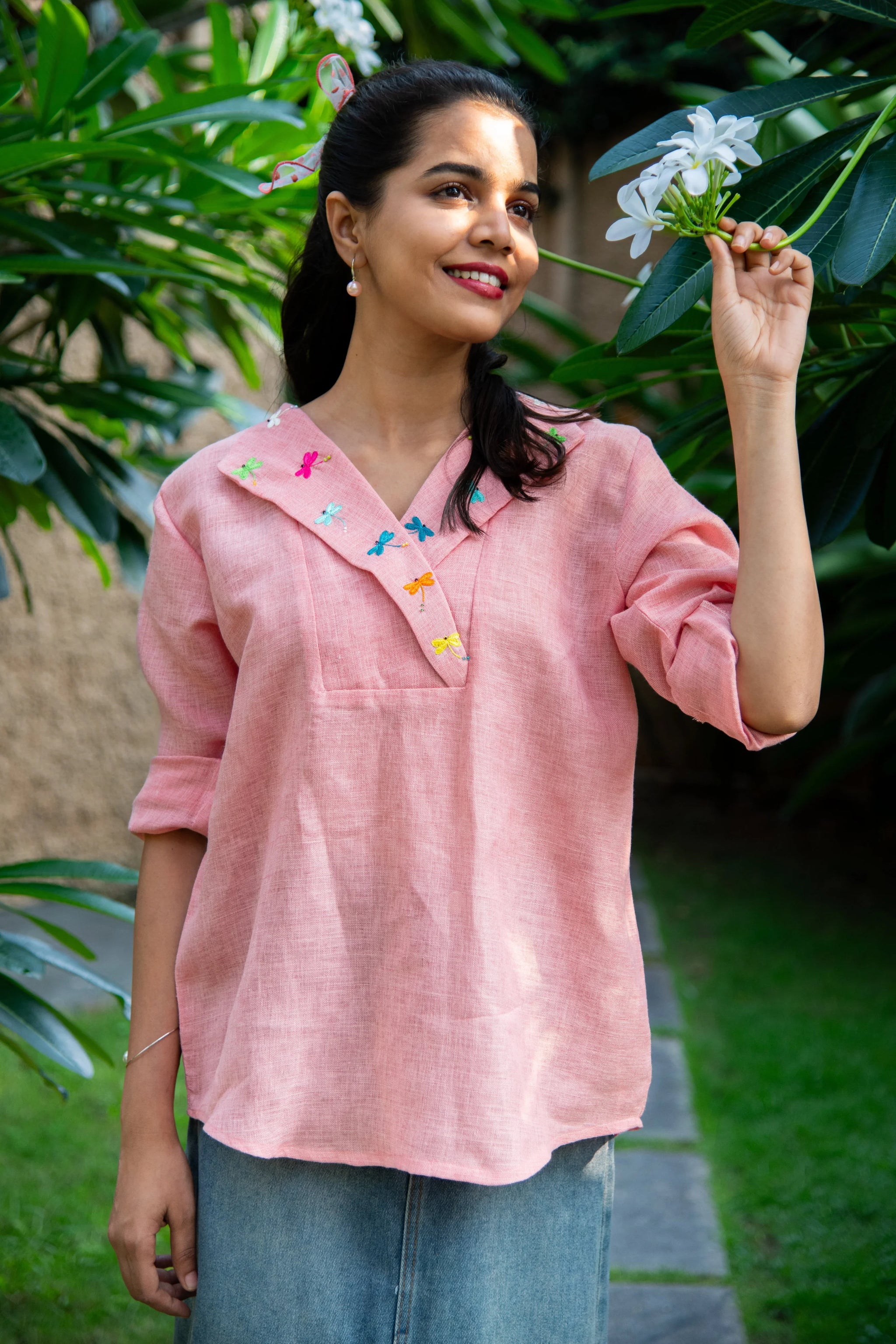 Woman in pink embroidered top with dragonfly detail and denim skirt in garden setting