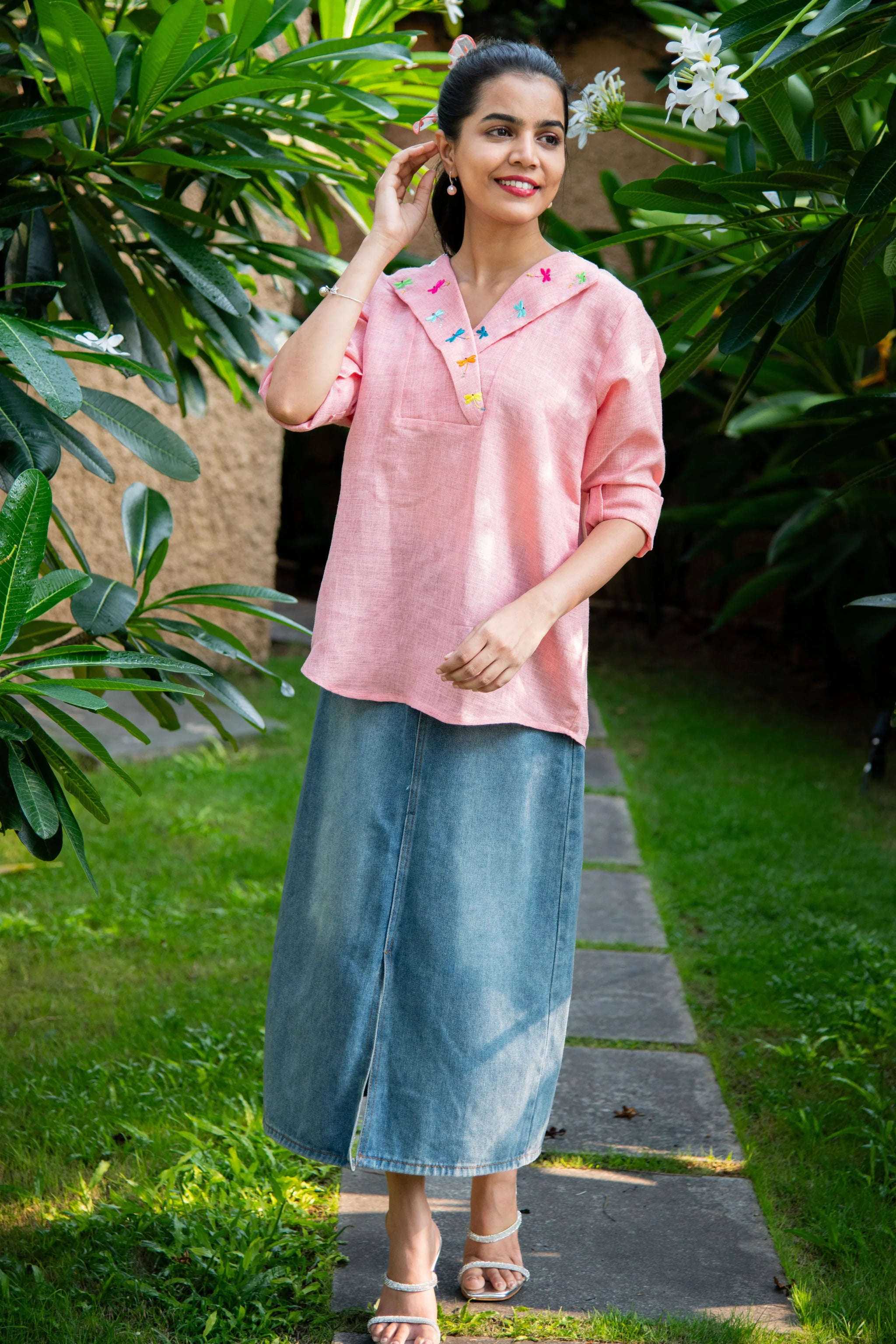 Woman in a pink embroidered top with long denim skirt standing in a garden