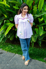 Woman in a lilac puff sleeve embroidered shirt and blue jeans standing outdoors with greenery