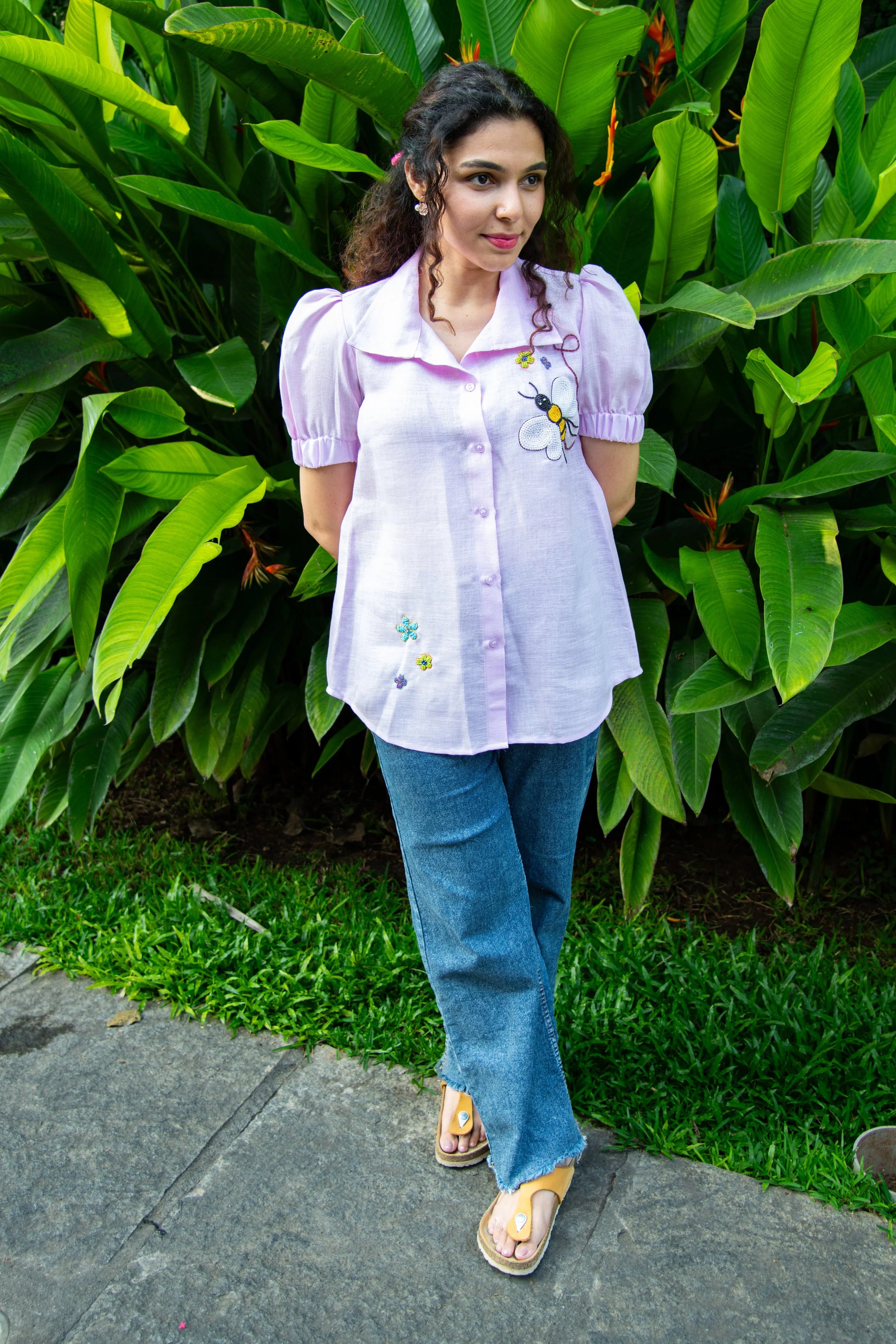 Woman in a lilac puff sleeve embroidered shirt and blue jeans standing outdoors with greenery