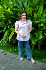 Woman in a light purple embroidered blouse and blue jeans standing outdoors by green plants