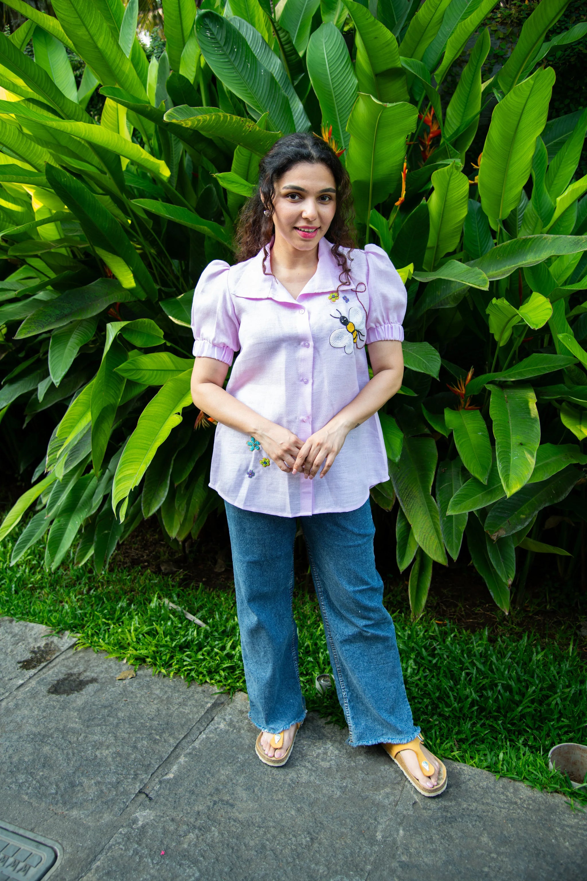 Woman in a light purple embroidered blouse and blue jeans standing outdoors by green plants