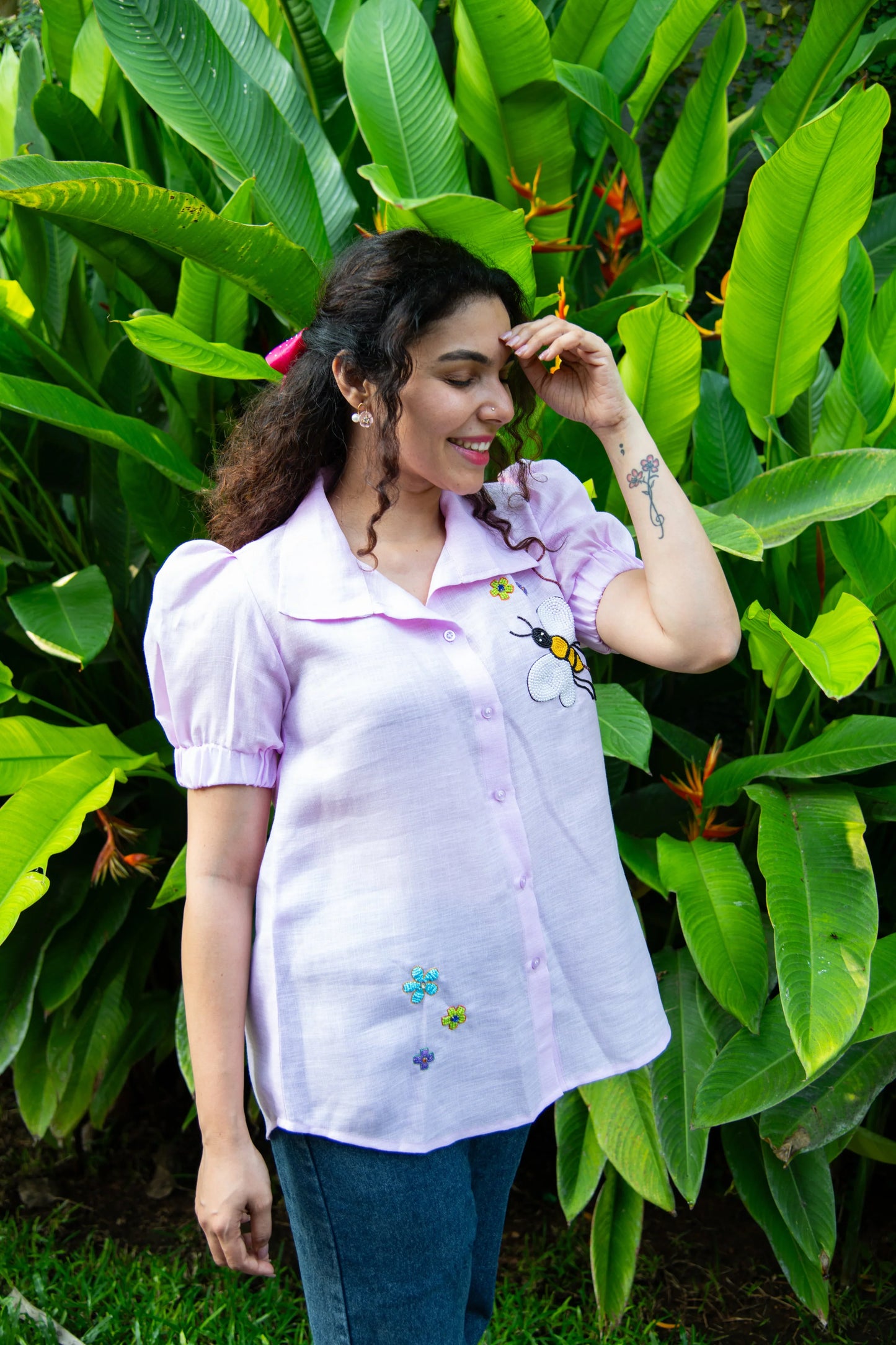 Woman in a lavender embroidered blouse and jeans standing outdoors by tropical plants