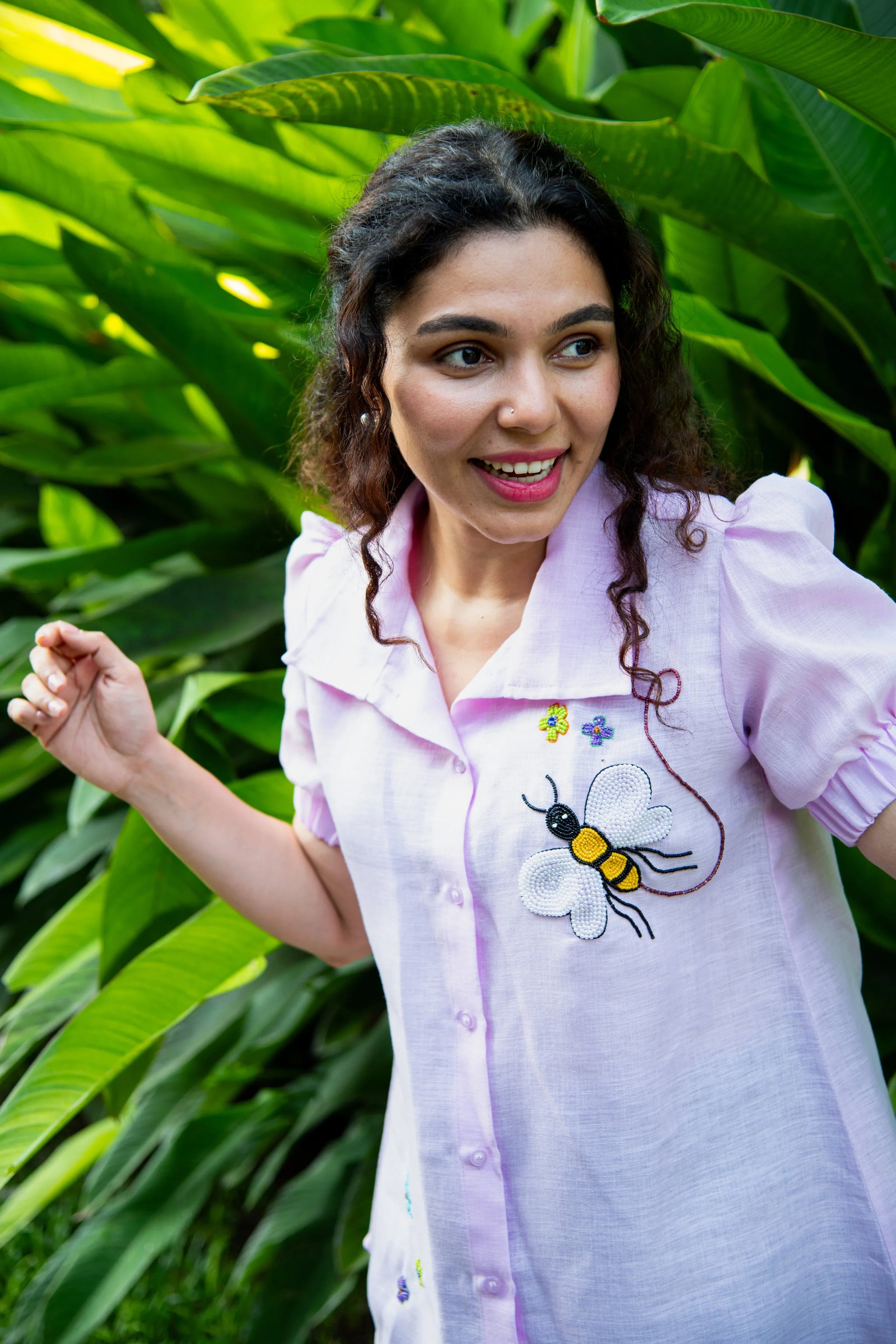 Woman in a light purple shirt with bee embroidery, standing by green tropical plants