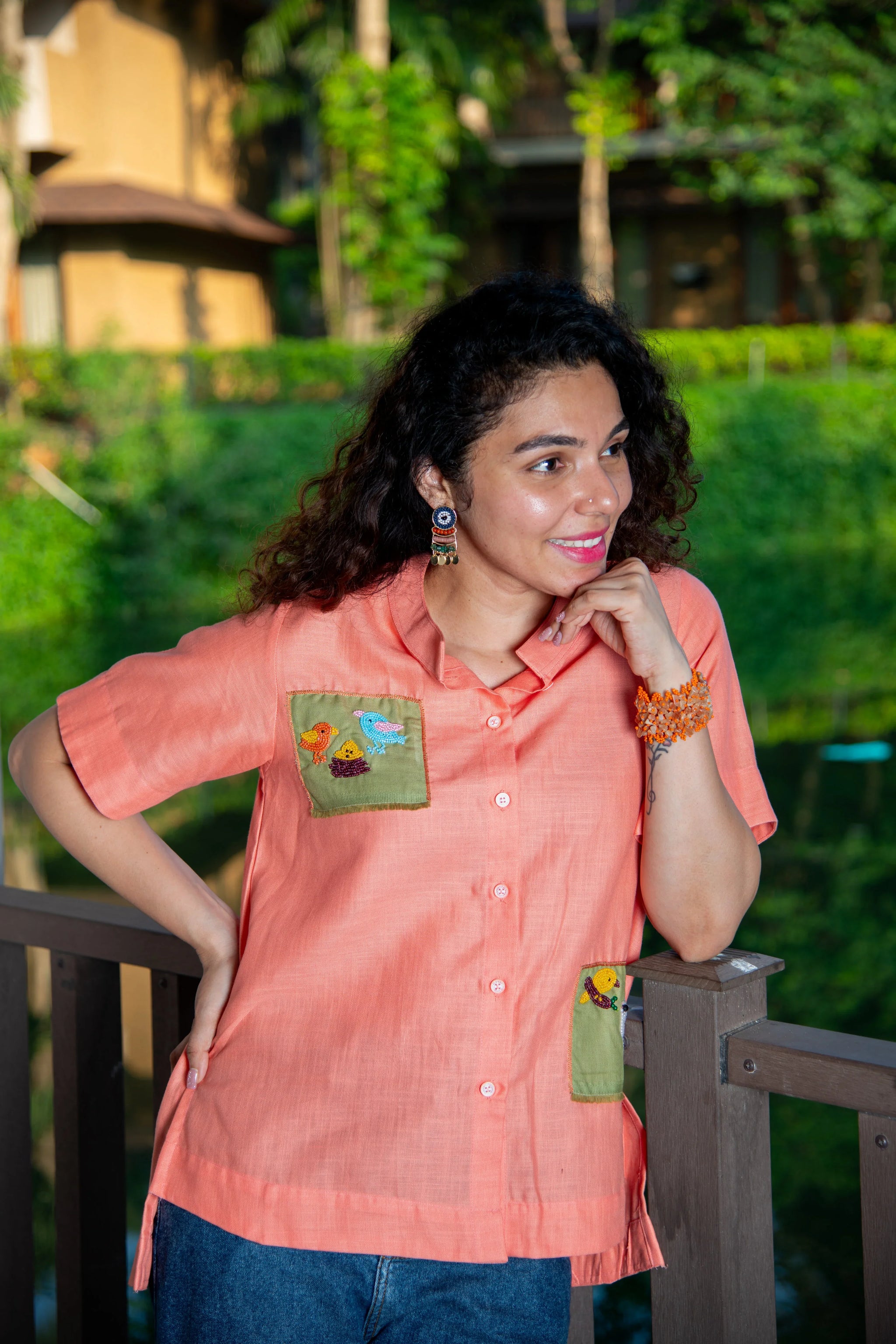 Woman in peach short-sleeve shirt with bird embroidery, standing outdoors on a balcony