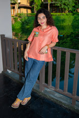 Woman in an orange shirt and blue jeans standing by a balcony with lush greenery in background