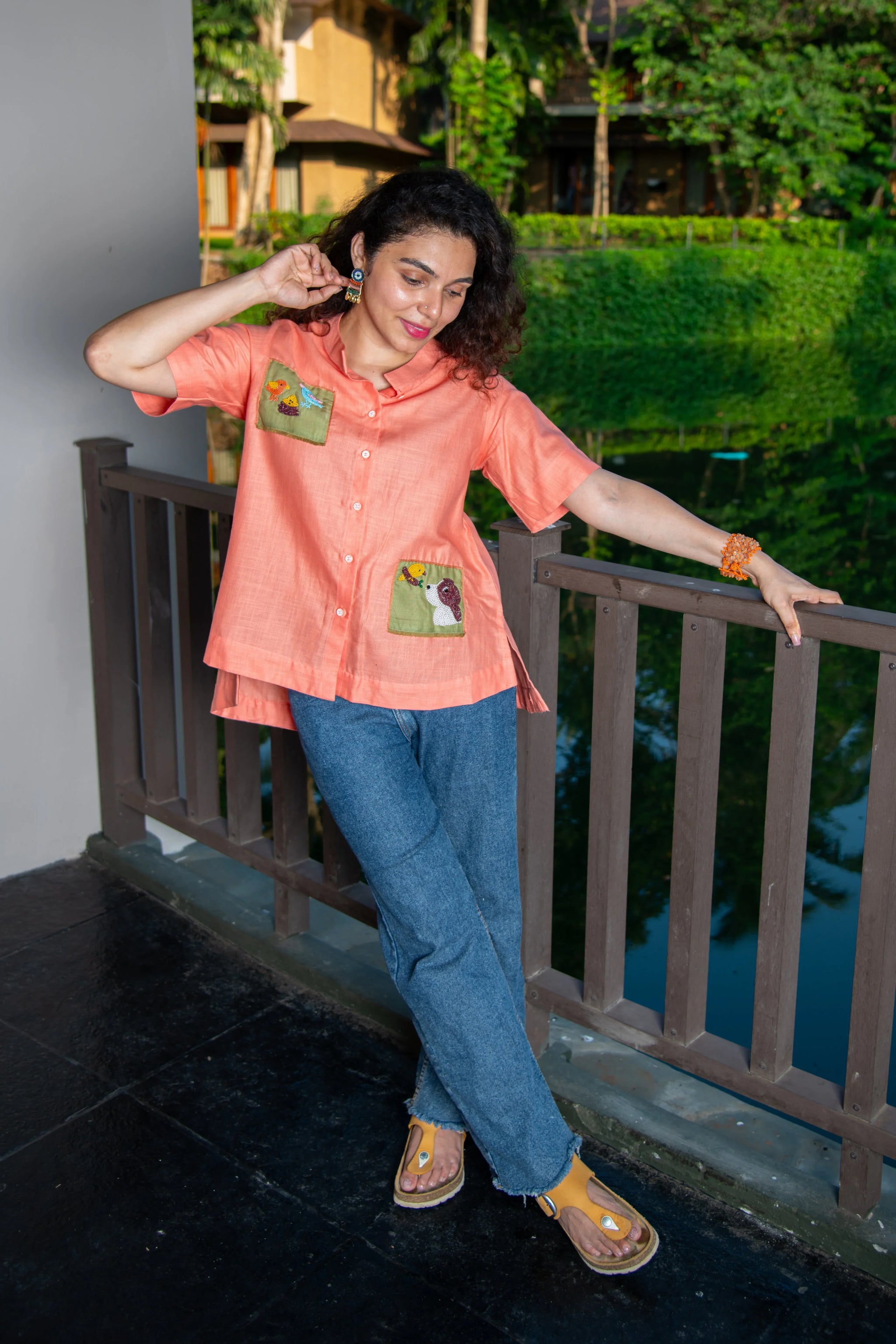 Woman in peach short-sleeve shirt with embroidered pockets and blue jeans on balcony