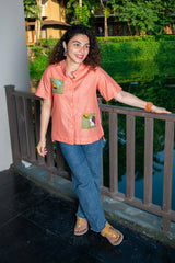 Woman in peach short sleeve shirt with embroidered pockets, blue jeans, and sandals outdoors