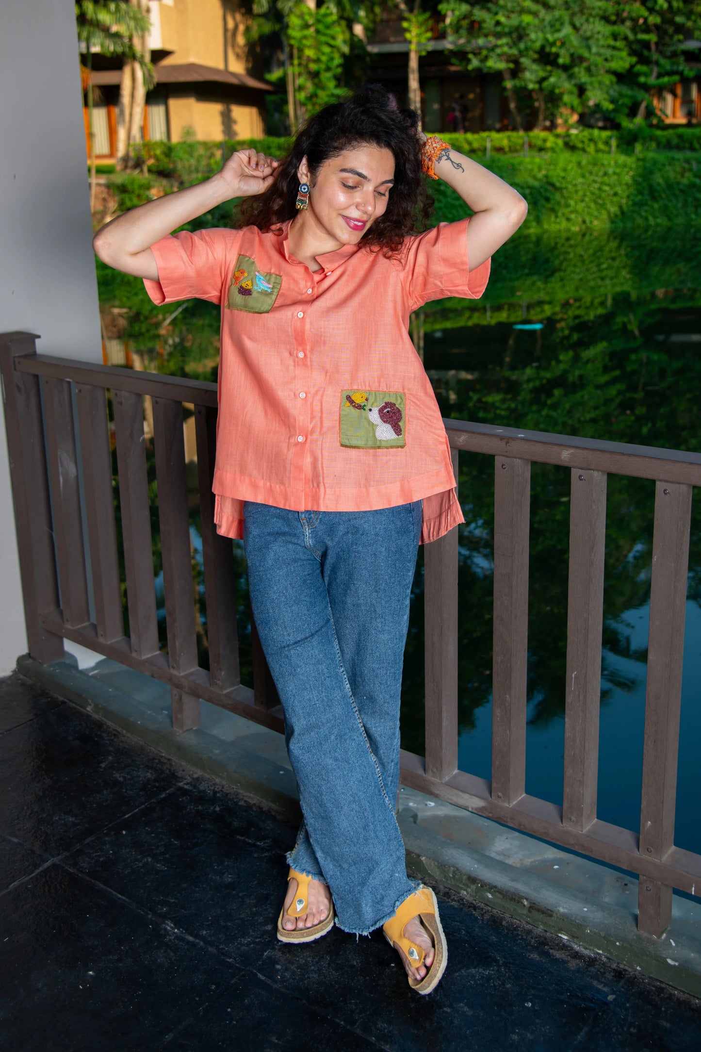 Woman in an orange shirt with embroidered pockets and blue jeans, standing on a balcony