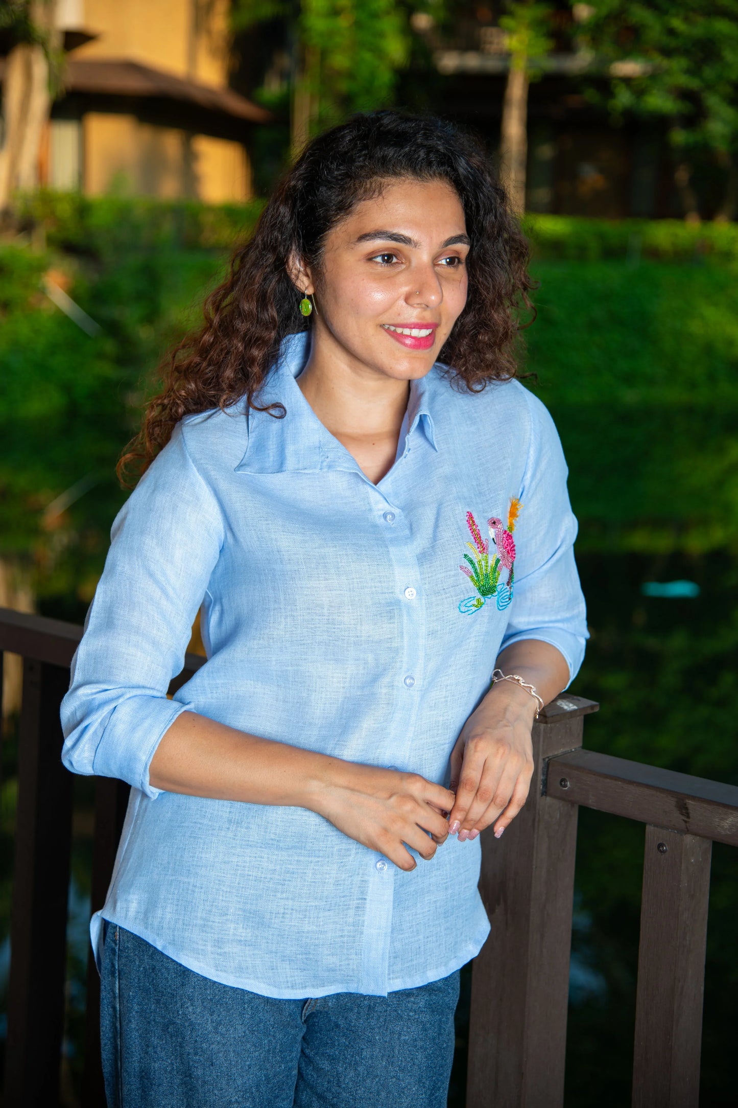 Woman in light blue embroidered shirt and jeans standing outdoors by a wooden railing