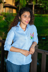 Woman in light blue embroidered shirt and jeans standing outdoors by a wooden railing