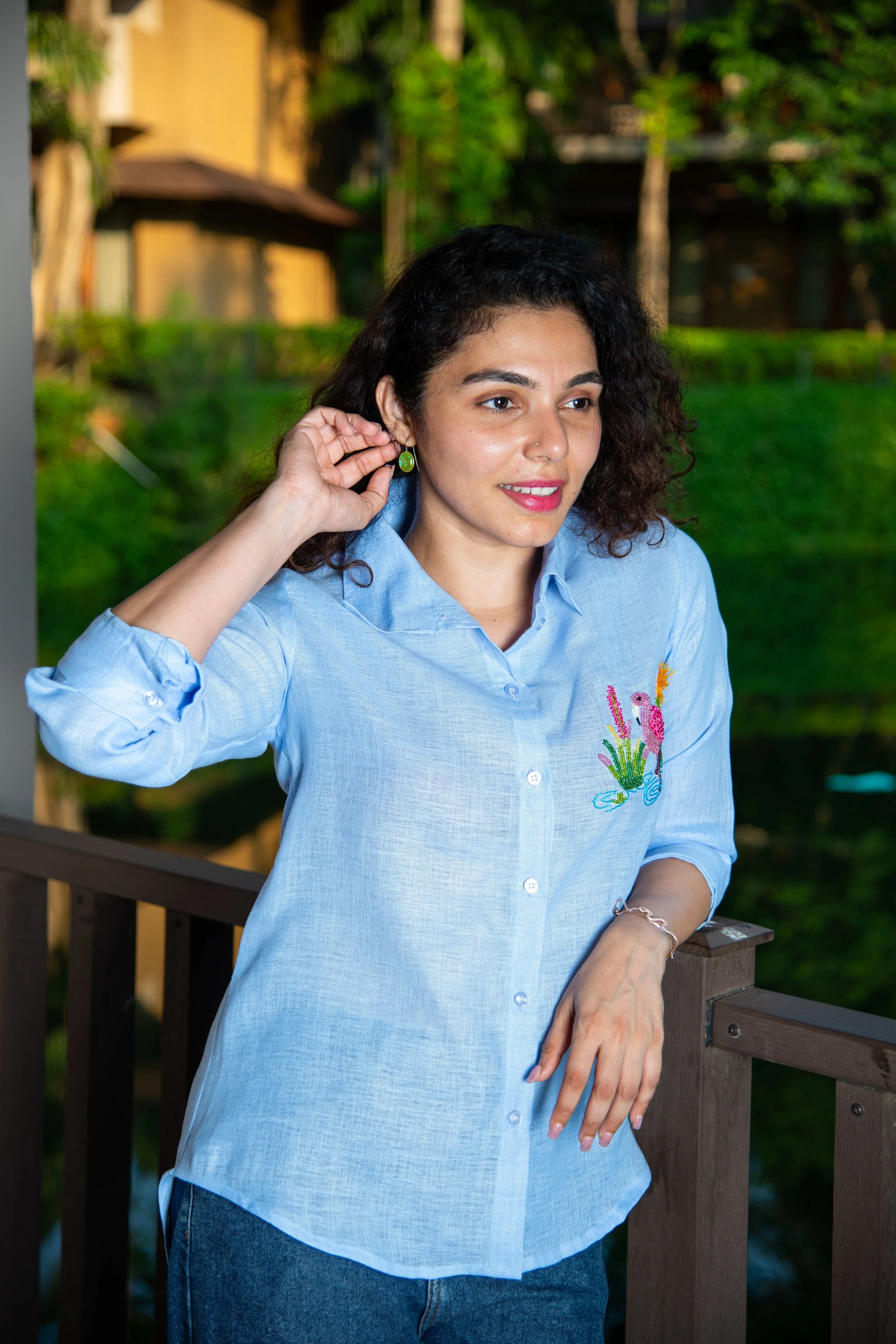 Woman in light blue embroidered shirt and jeans standing outdoors by a wooden railing