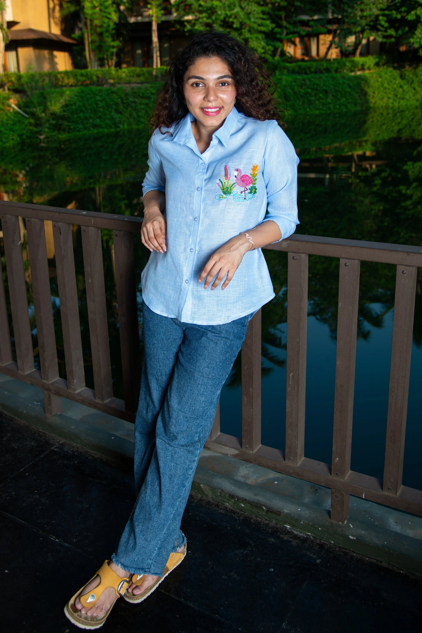 Woman in a light blue embroidered shirt and denim jeans, standing on a wooden bridge.