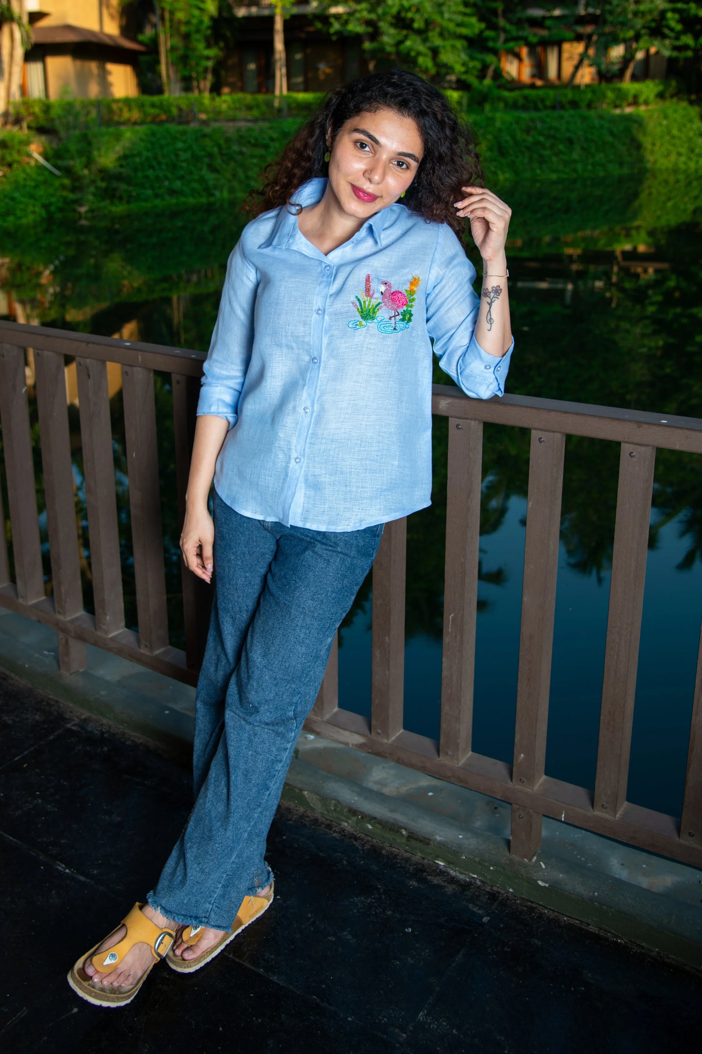 Woman in light blue embroidered shirt and blue jeans standing outdoors by a railing