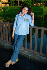 Woman in light blue embroidered shirt and blue jeans standing outdoors by a railing