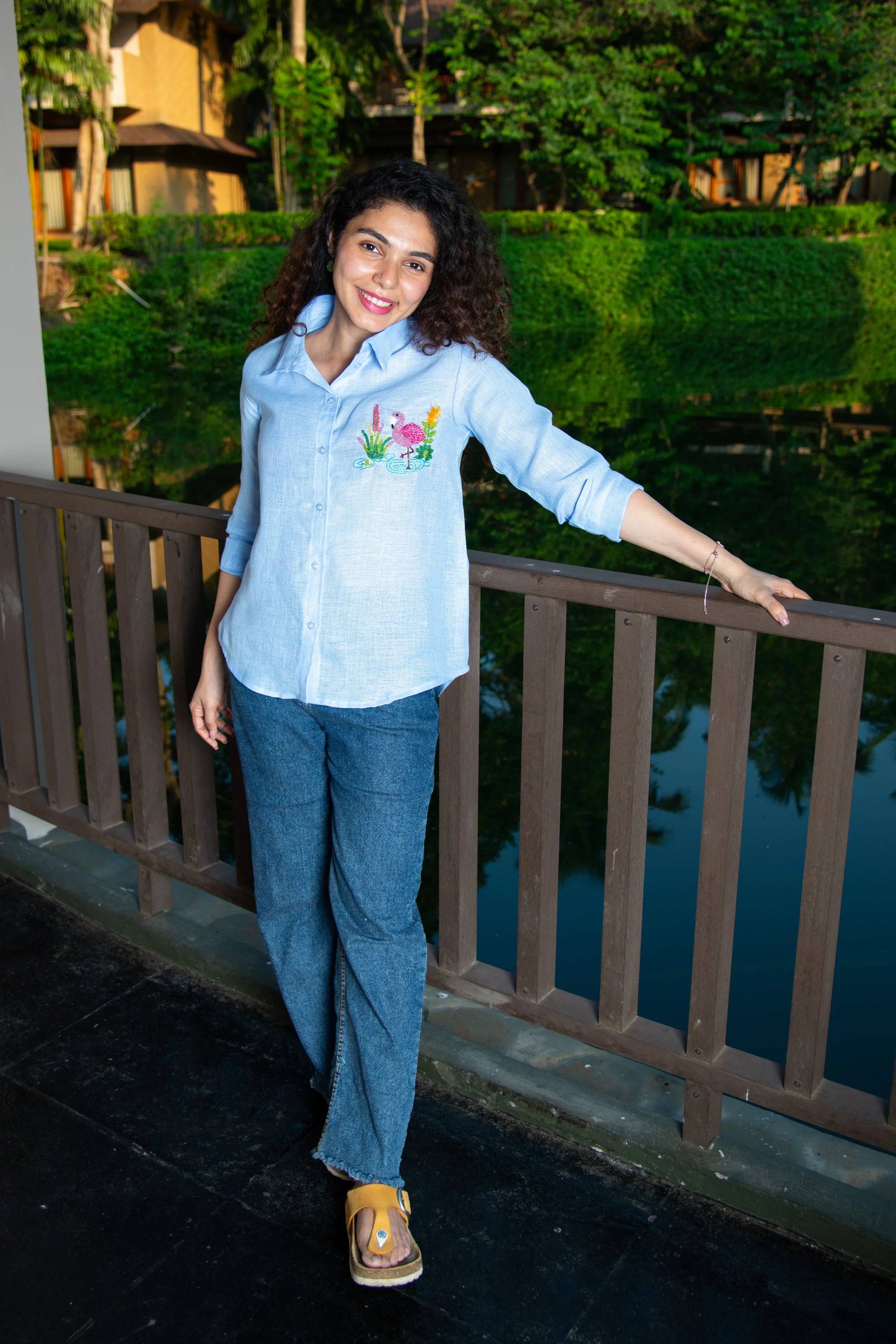 Woman in a light blue embroidered shirt and jeans posing on a wooden bridge outdoors