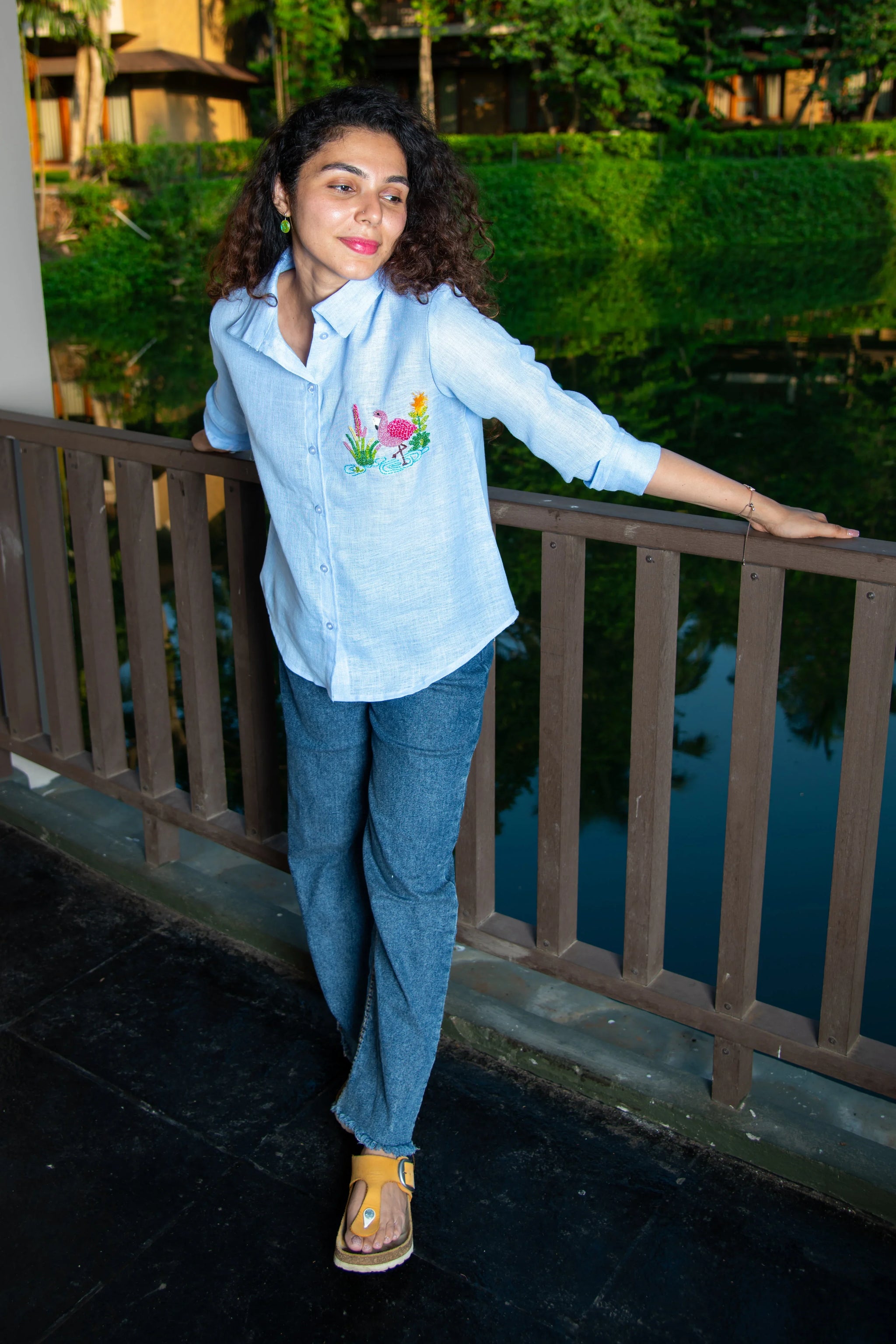 Woman in a light blue embroidered shirt and blue pants standing by a wooden railing outdoors