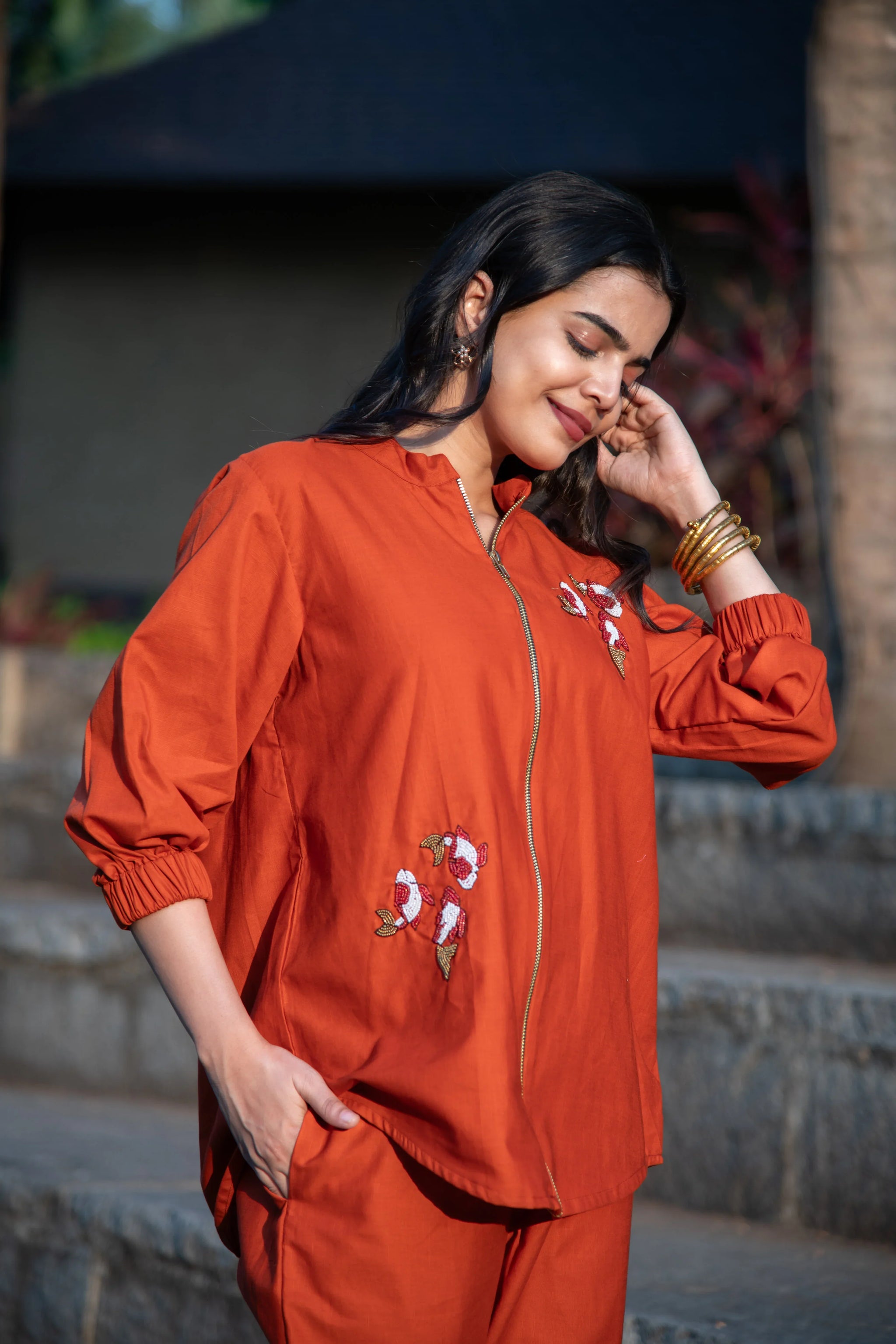 Woman in burnt orange embroidered co-ord set with zipper and gold bangles outdoors