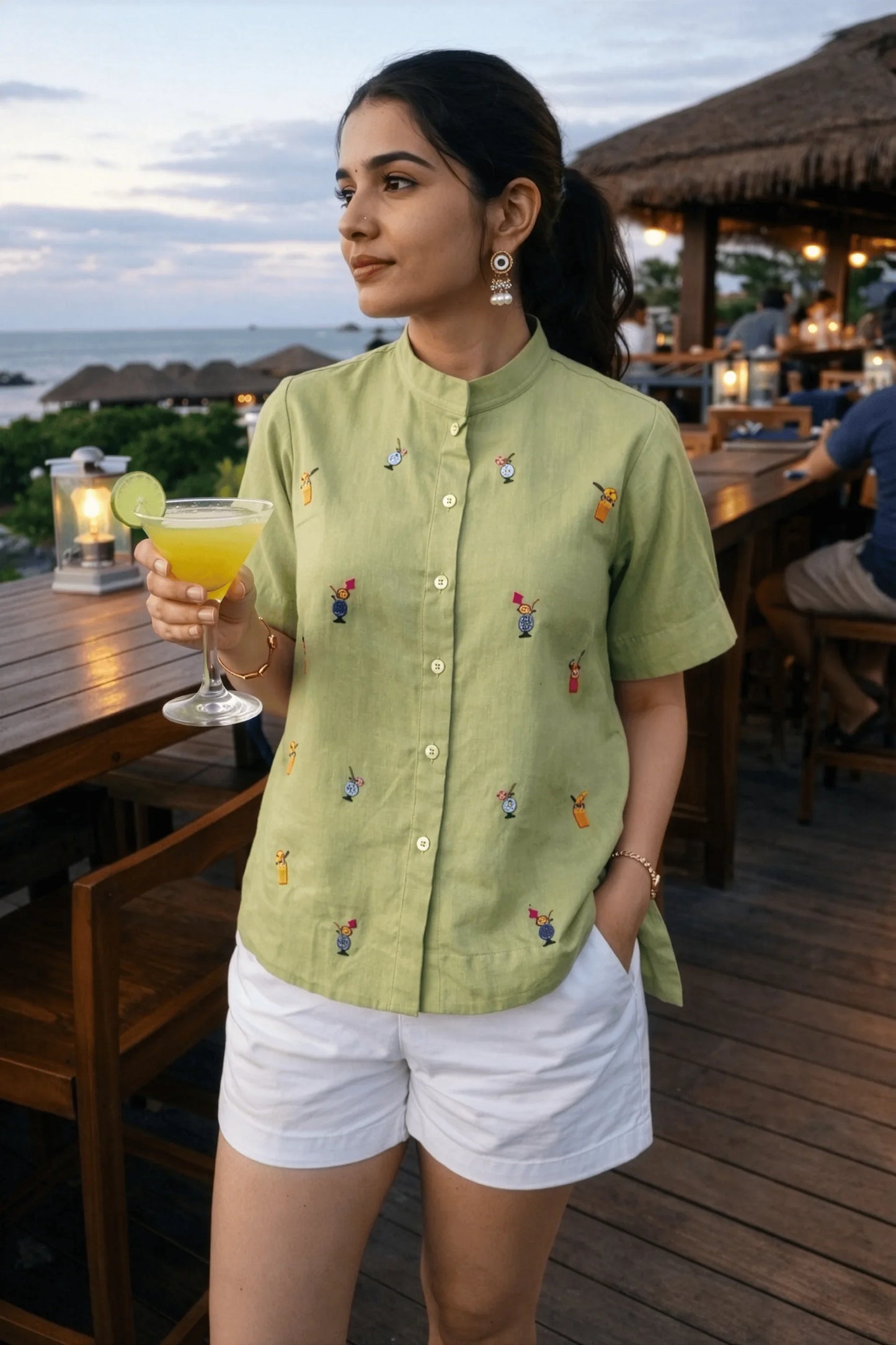 Woman in green embroidered shirt and white shorts holding cocktail at beachside bar