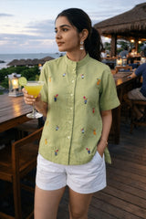 Woman in green embroidered shirt and white shorts holding cocktail at beachside bar