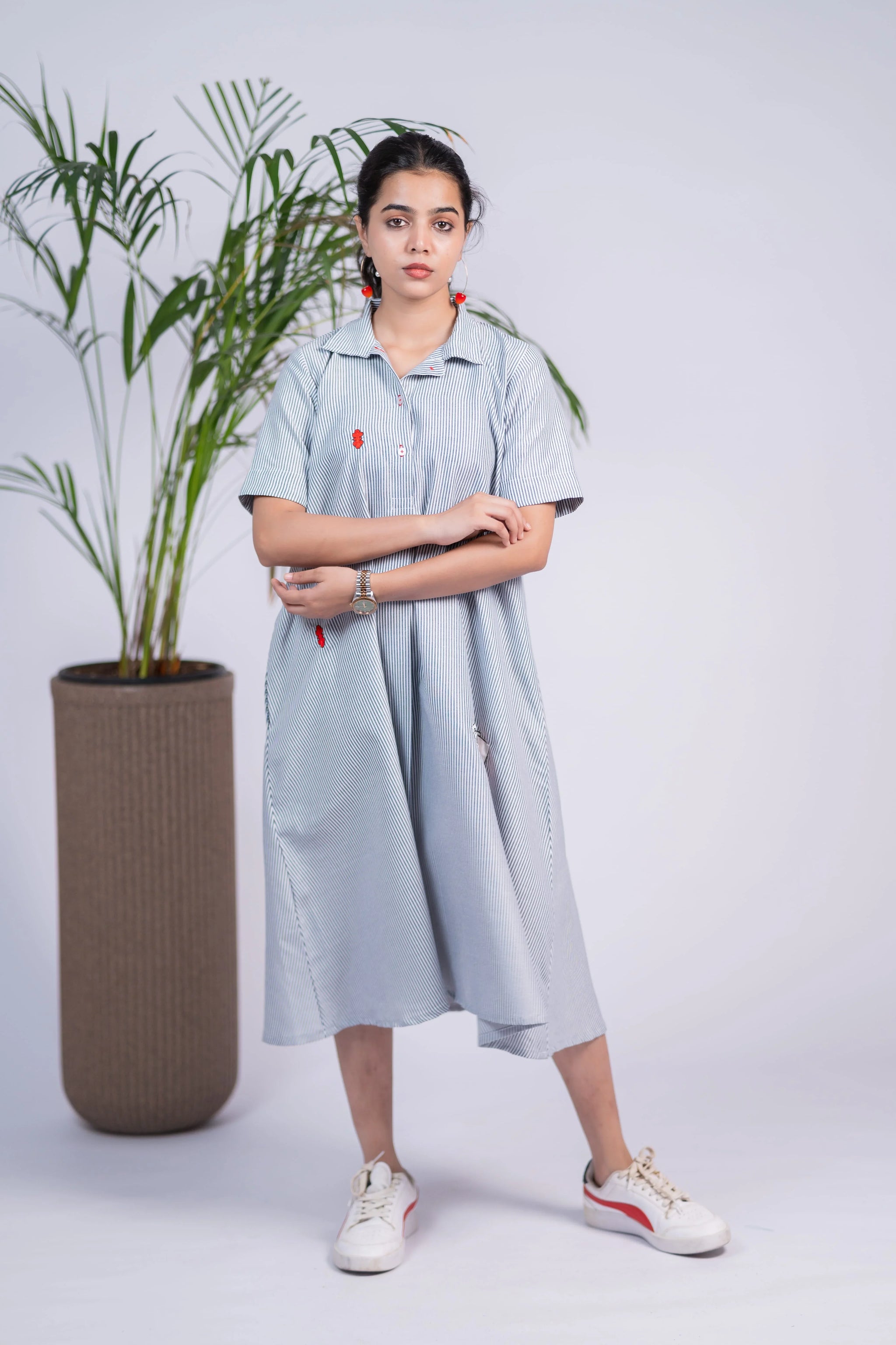 Woman in a light blue striped midi shirt dress with red accents, casual sneakers, indoor plant backdrop