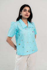 Light blue short sleeve shirt with colorful charm embellishments, paired with white pants for a trendy everyday look.