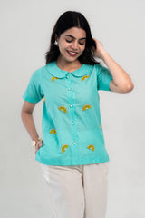 Turquoise lemon-embroidered shirt with pearl buttons, paired with cream pants for a trendy look.