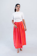 Woman in white ribbed top and vibrant orange-pink striped pleated midi skirt, styled with clear heels and cherry earrings.