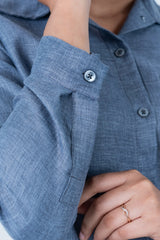 Blue linen shirt with buttoned cuffs and subtle texture, paired with a delicate ring