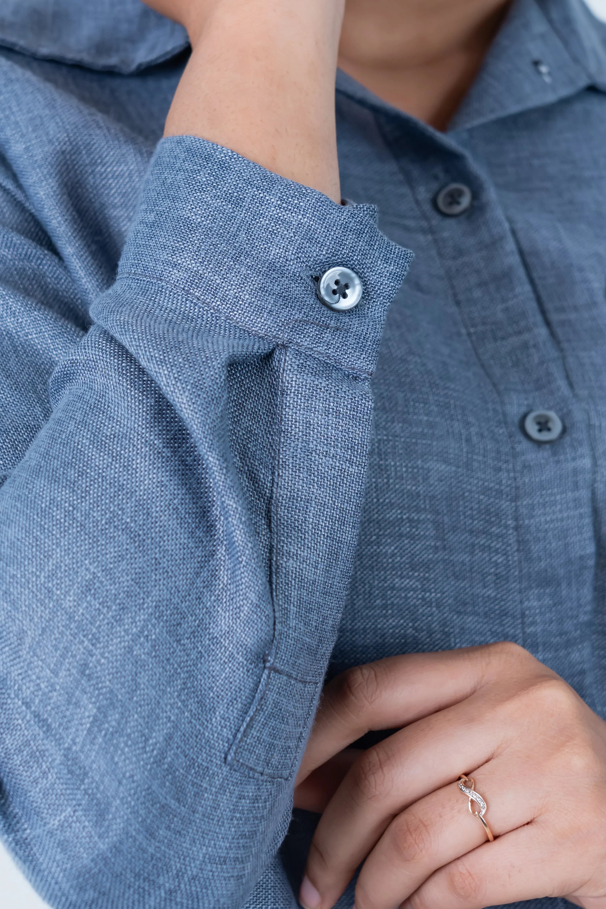 Blue linen shirt with buttoned cuffs and subtle texture, paired with a delicate ring