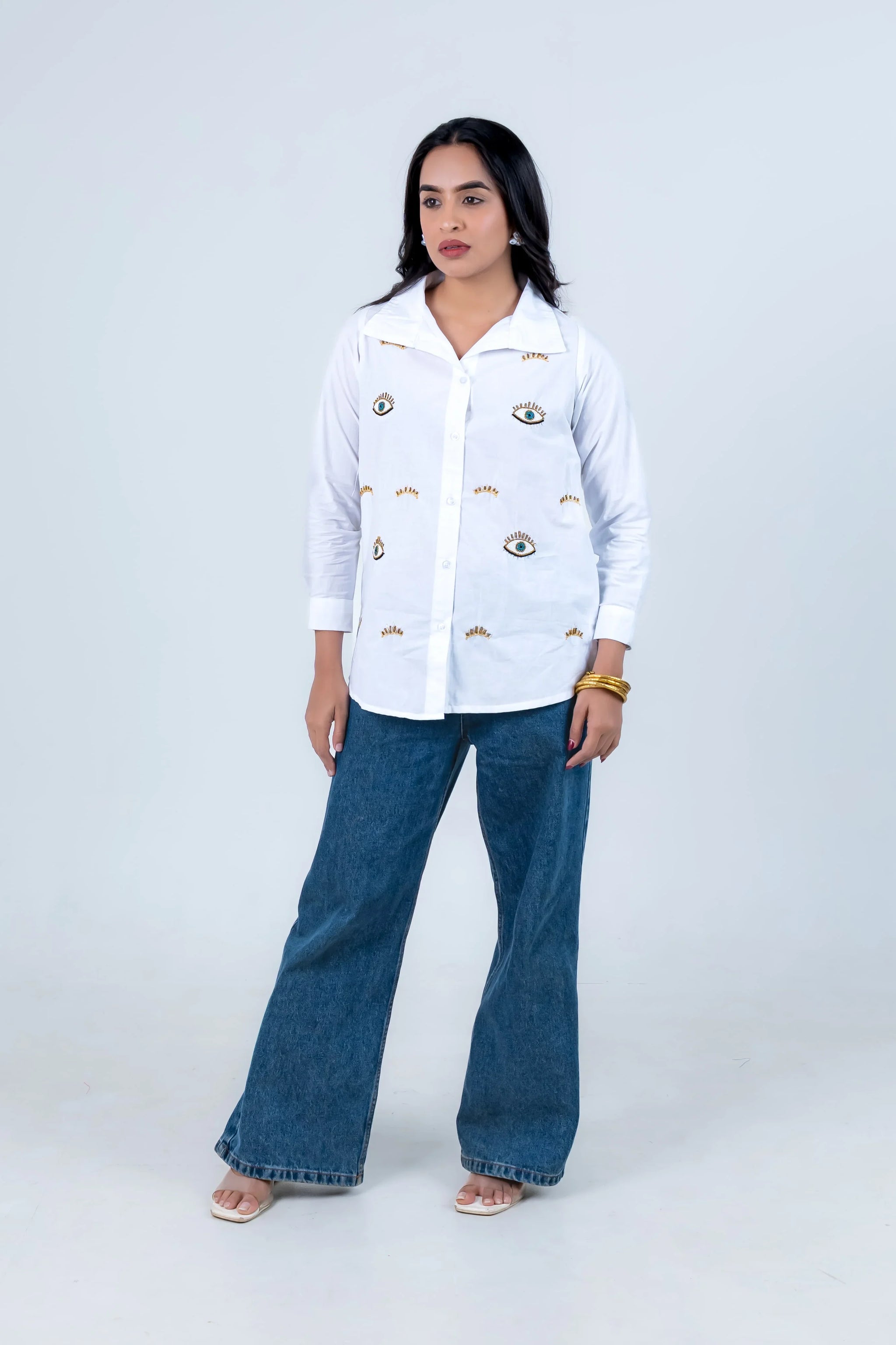 White button-down shirt with eye embroidery, paired with blue wide-leg jeans and sandals.