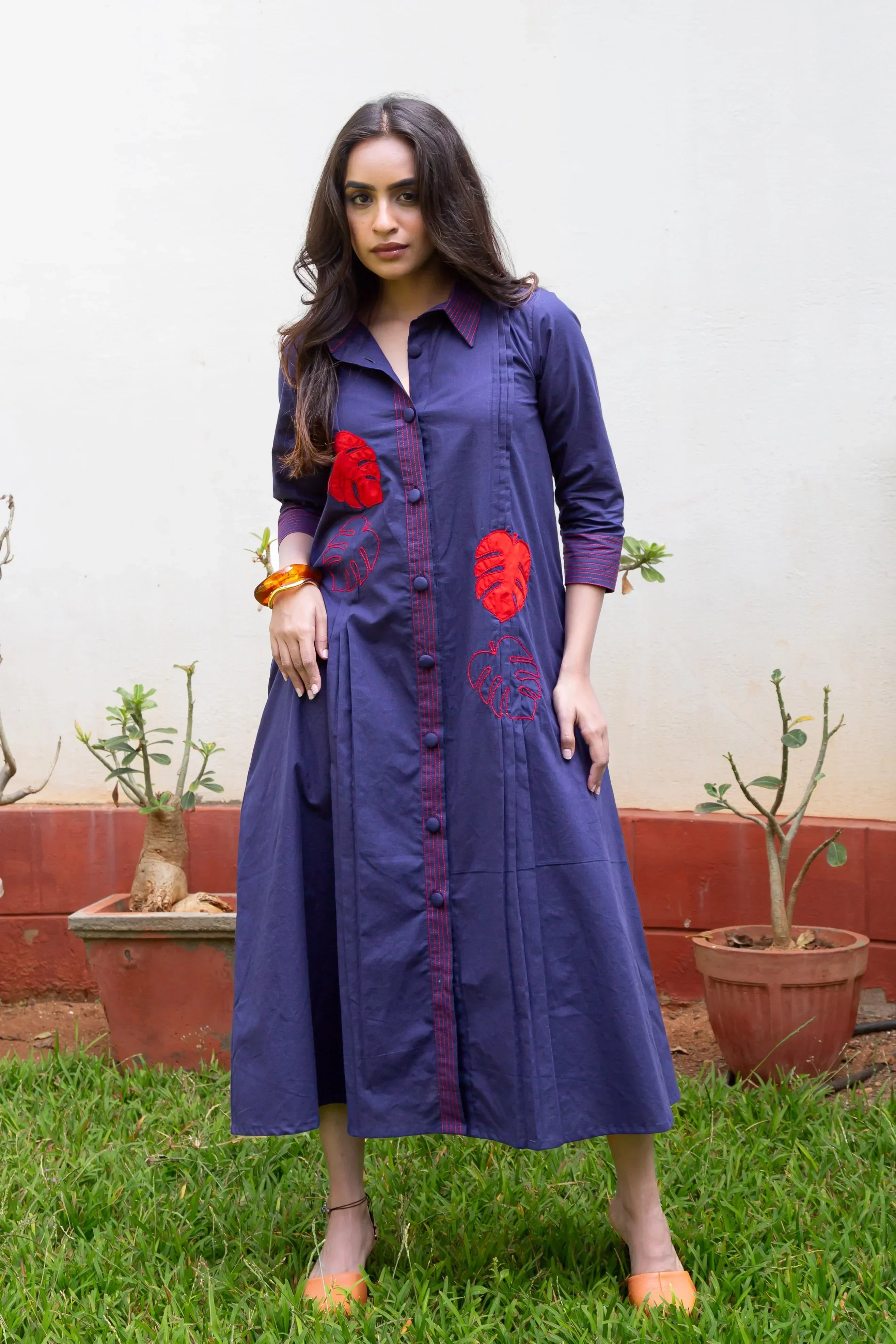 Woman wearing a navy blue embroidered dress with red leaf patterns and orange shoes standing on grass