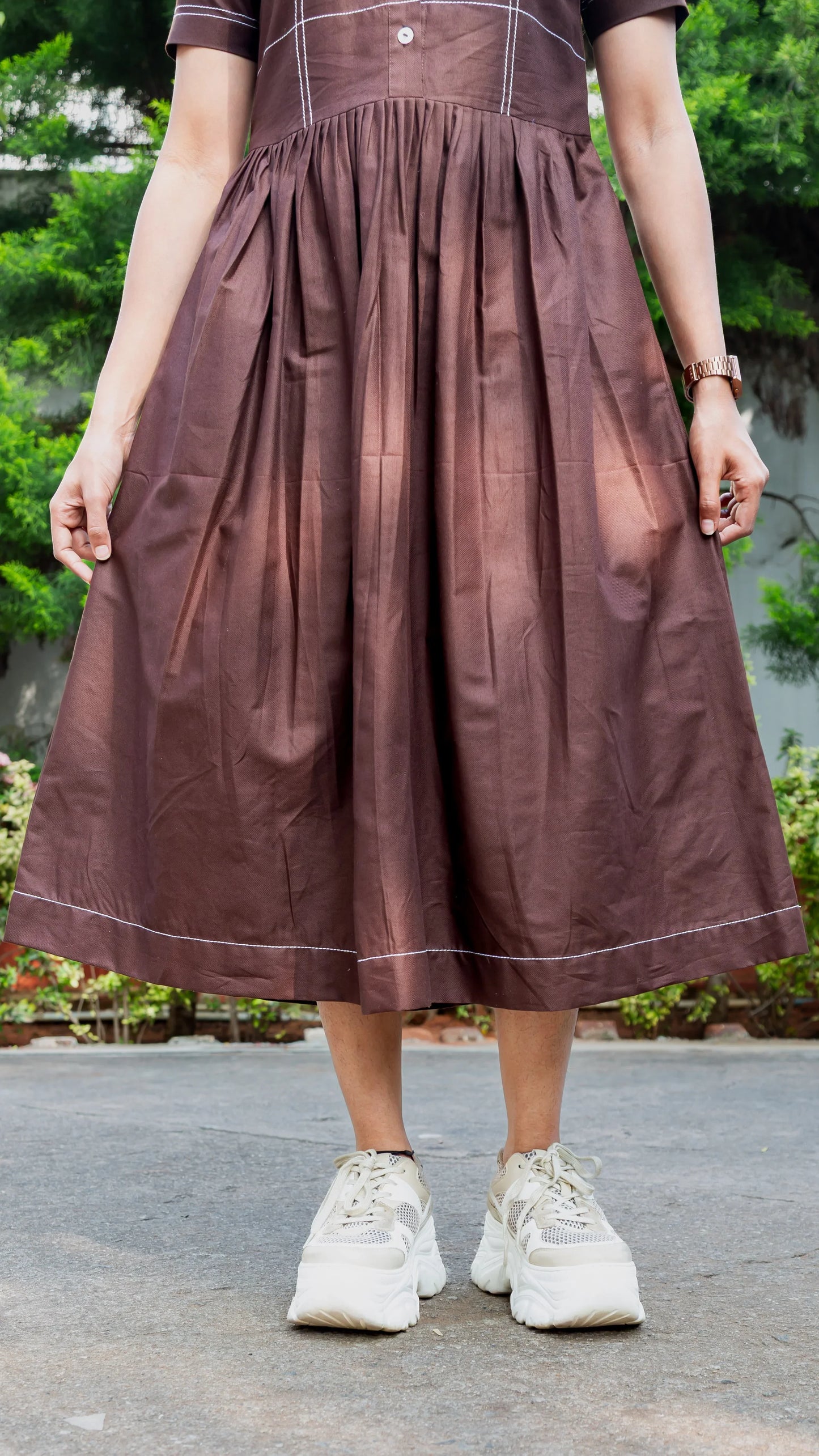 Brown pleated midi dress with white stitching, paired with chunky white sneakers for a trendy look.