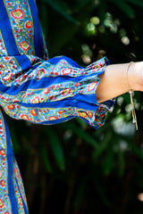 Blue floral dress sleeve with ruffled detail and gold bracelet against green foliage