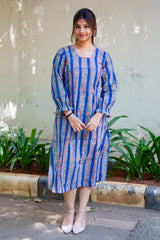 Woman in blue floral print dress with long sleeves, standing outdoors near green plants