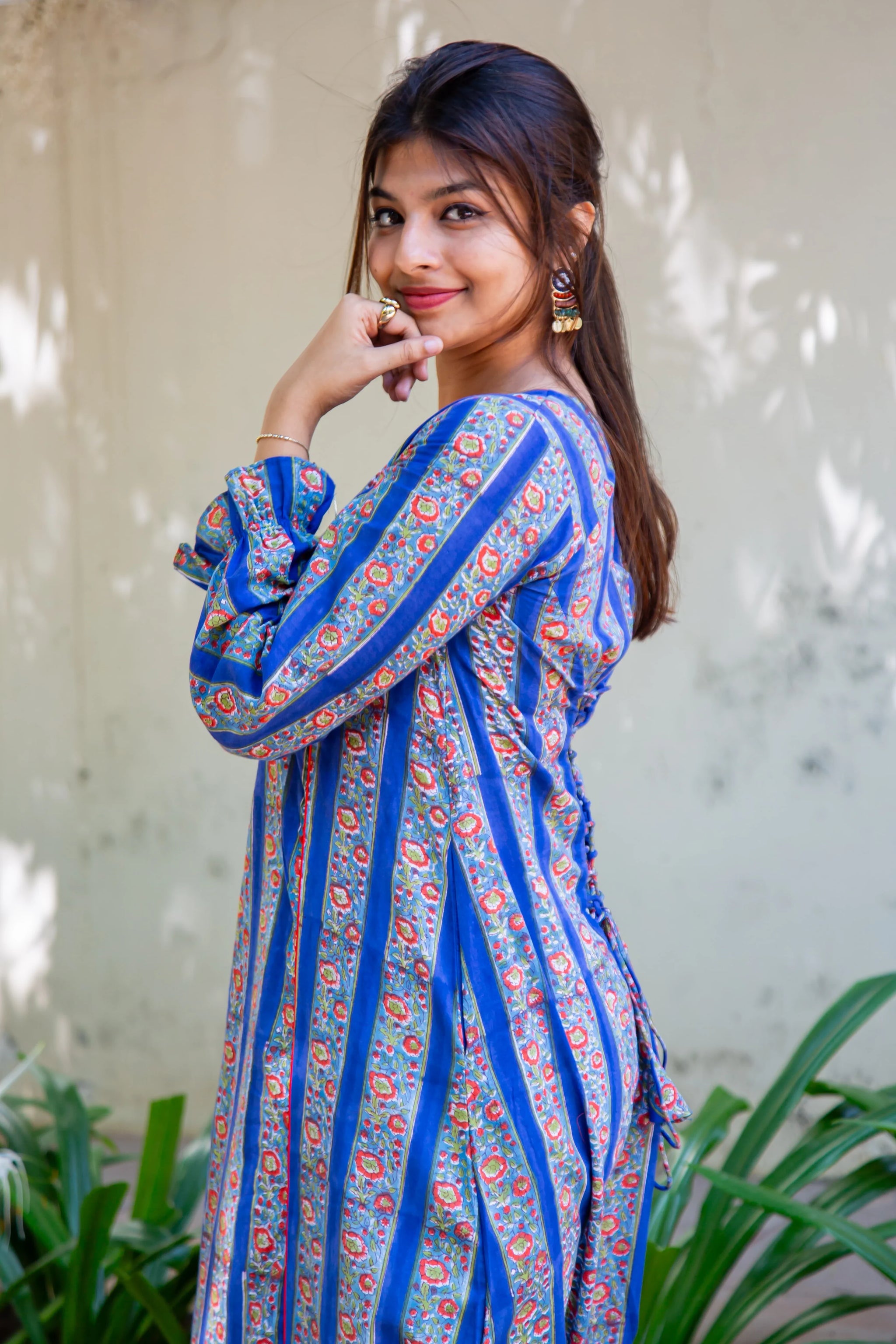 Woman in blue floral dress with ruffled sleeves, posing outdoors near greenery