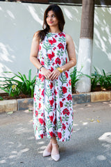 Woman in white sleeveless dress with red floral print, standing outdoors near green plants and tree.